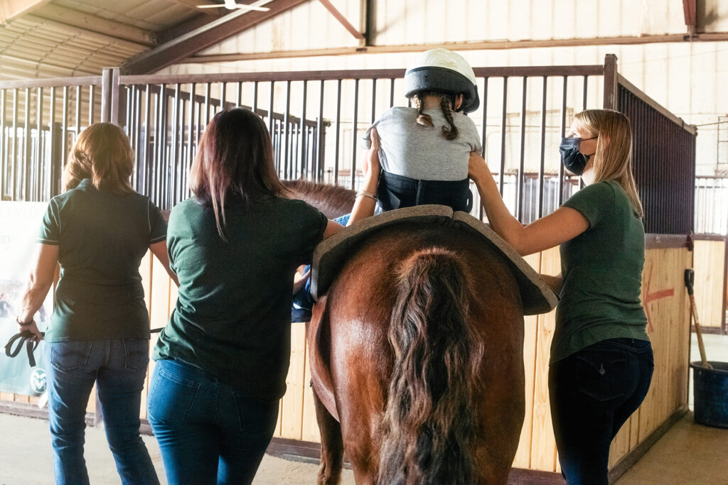 Three people assist a girl in a helmet on a horse.