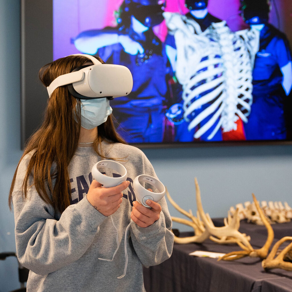 Girl in a sweatshirt uses a virtual reality headset and hand-held remotes.