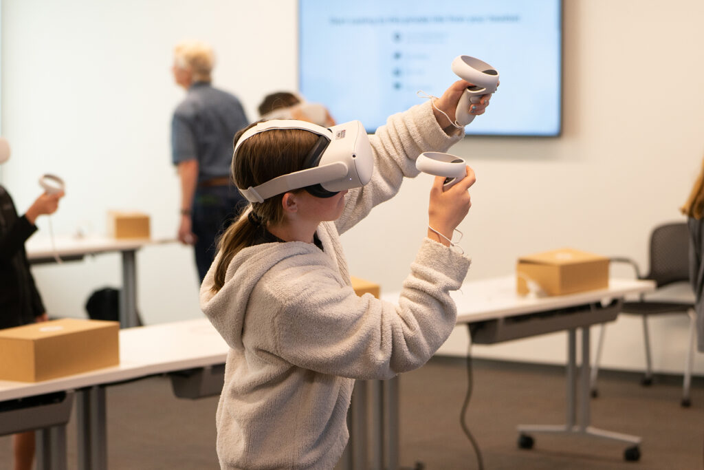 A child in a white sweatshirt uses a virtual reality headset and corresponding hand remotes.