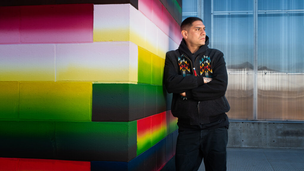 A man in a black hoodie looking away from the camera leans against a brightly colored wall.