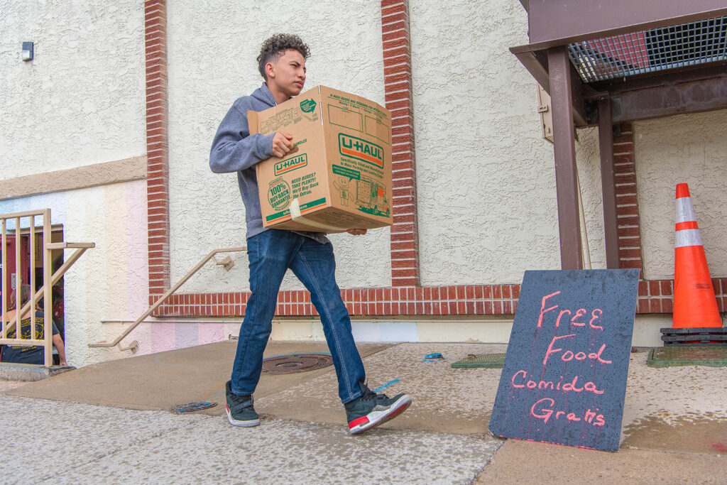 A person carries a cardboard box past a chalkboard sign that says free food / comida gratis