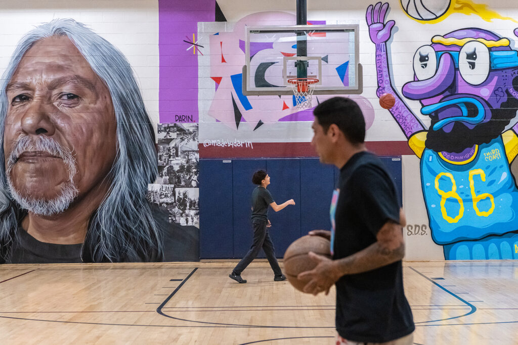 A man holds a basketball on a basketball court with a mural of an older man on the wall.