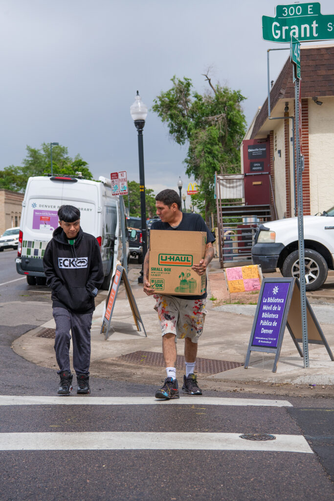 Two people cross the street in a crosswalk, one carrying a large cardboard box.