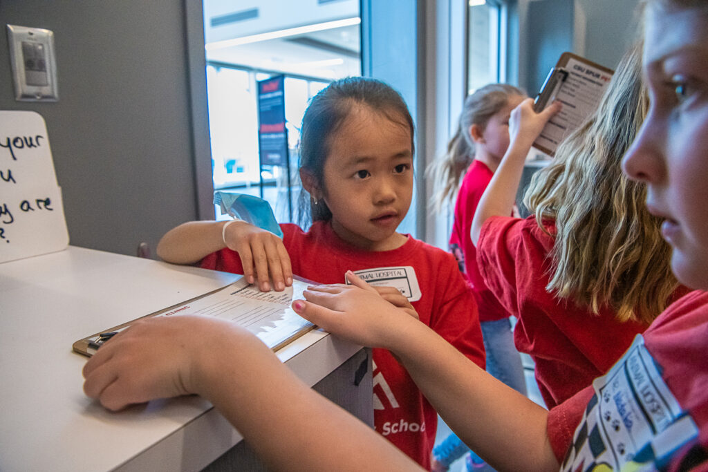 Children in red shirts write on a clipboard in a mock veterinary clinic.