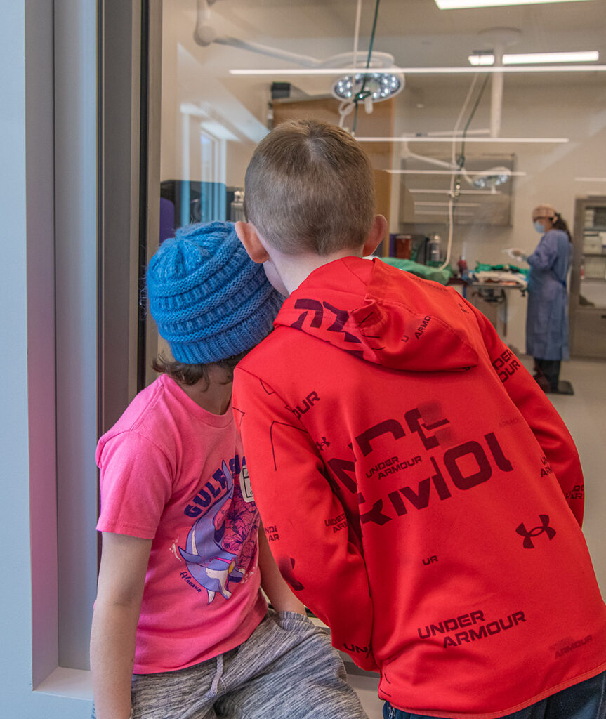 Two kids look through a large glass window at an animal surgery suite.