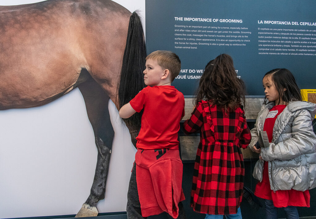 Three kids stand in front of an exhibit with a lifelike horse tail.