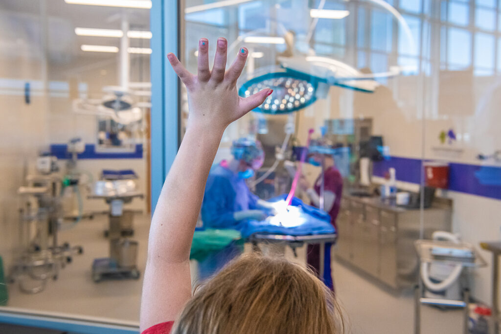 Child with hand in the air in front of a glass window looking into a surgery suite.