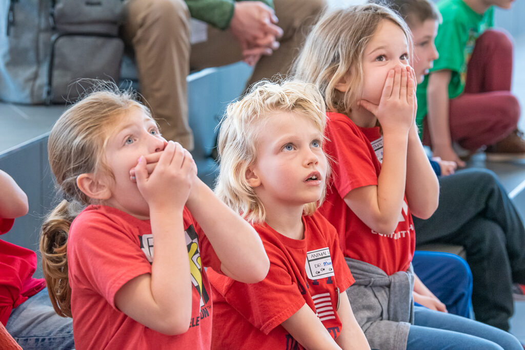 Three kids in red t-shirts looking shocked, two with hands over their mouths.