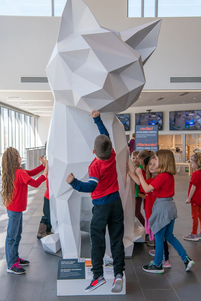 Kids in red shirts crowd around a large white cat statue.