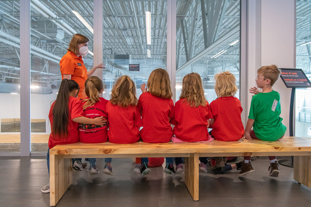 Kids in red shirts sit on a bench in front of a large glass window; a woman in a Spur shirt and headset stands to the left.