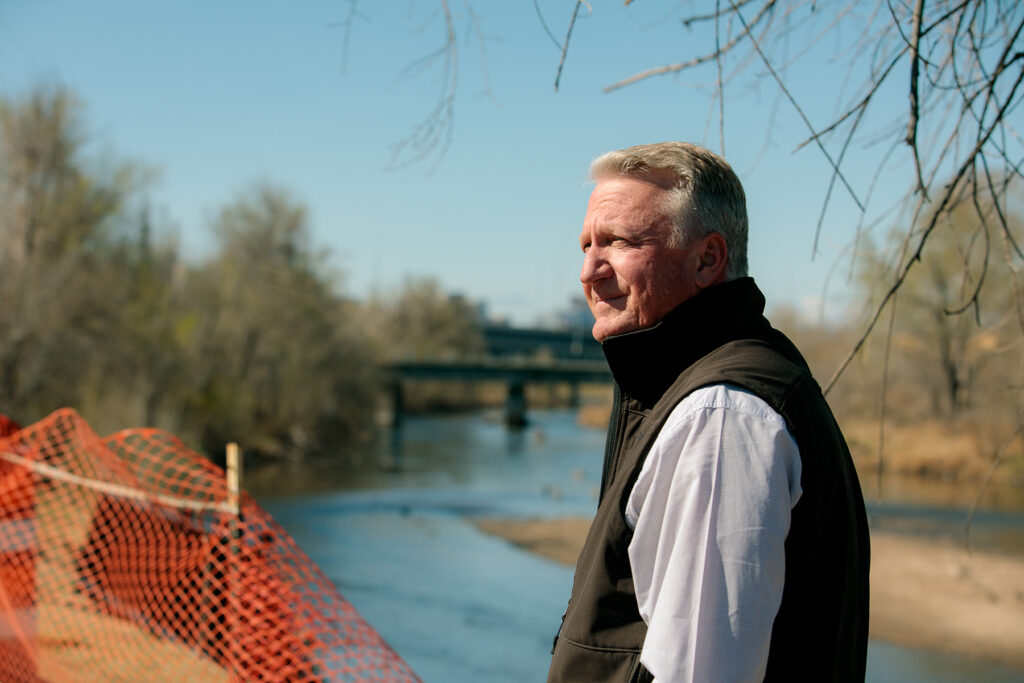 A person in a button up and a vest looks off to the side of the camera, standing in front of a river.