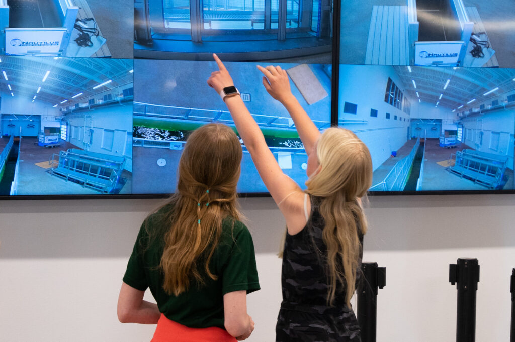 Two young people pointing up at a TV screen showing multiple images.