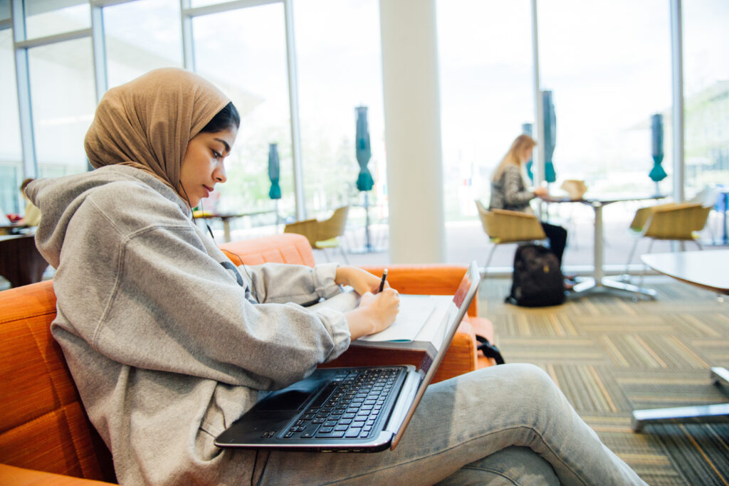 A student sits with a laptop and notebook in a common area.