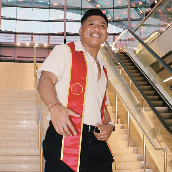 A man in a graduation sash stands on a staircase.