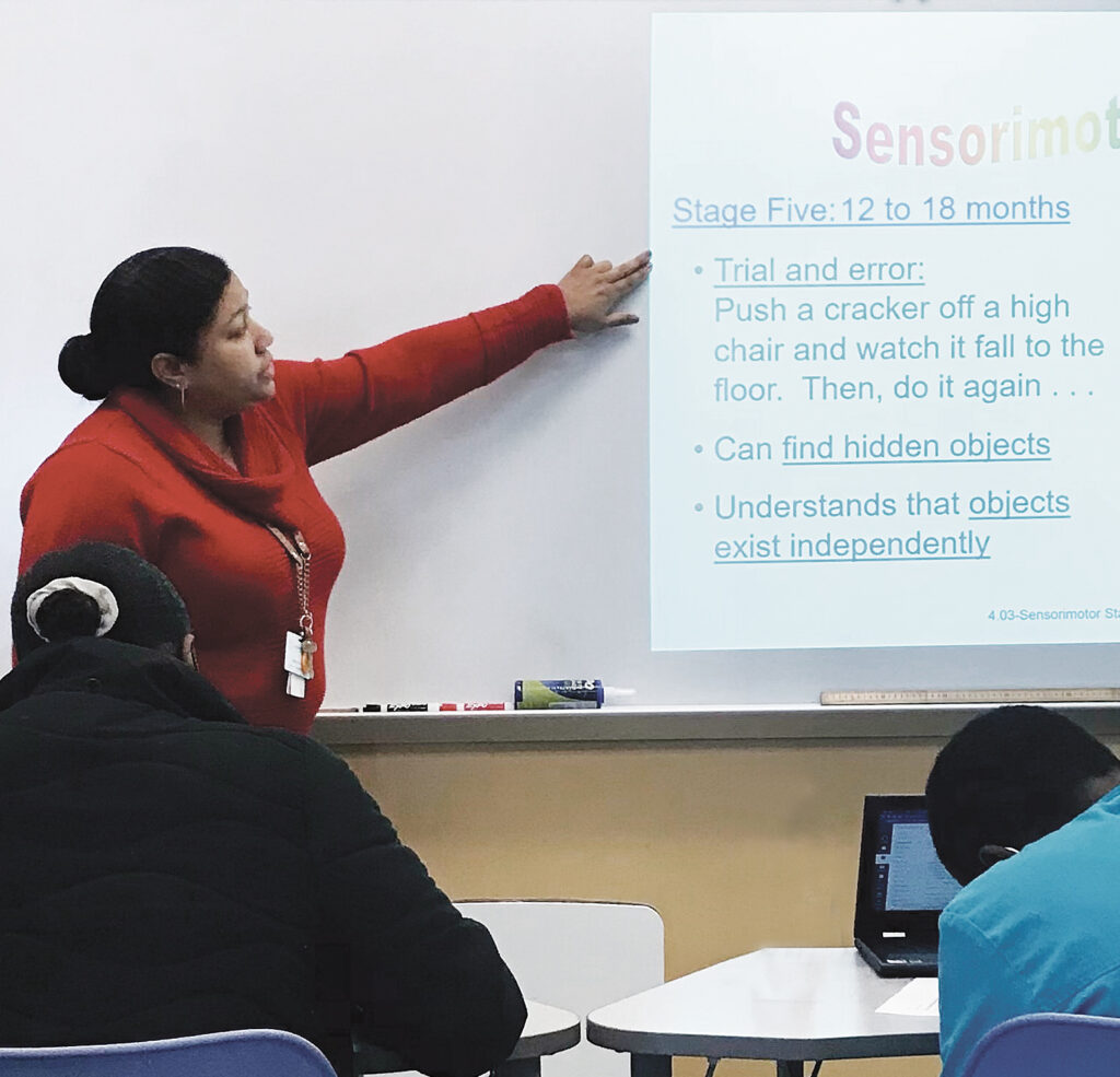 A woman in a red sweater points to something on a screen in front of a classroom.