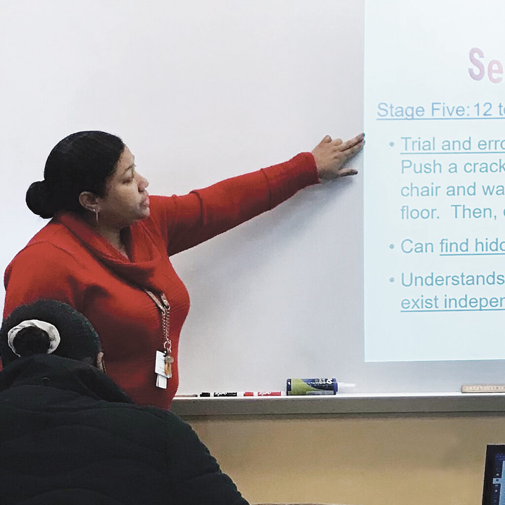 A woman in a red sweater points to something on a screen in front of a classroom.