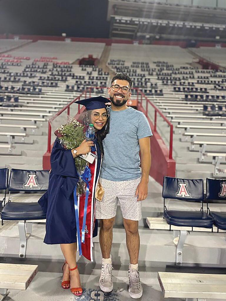 Two people stand together, one wearing graduation regalia and holding flowers.