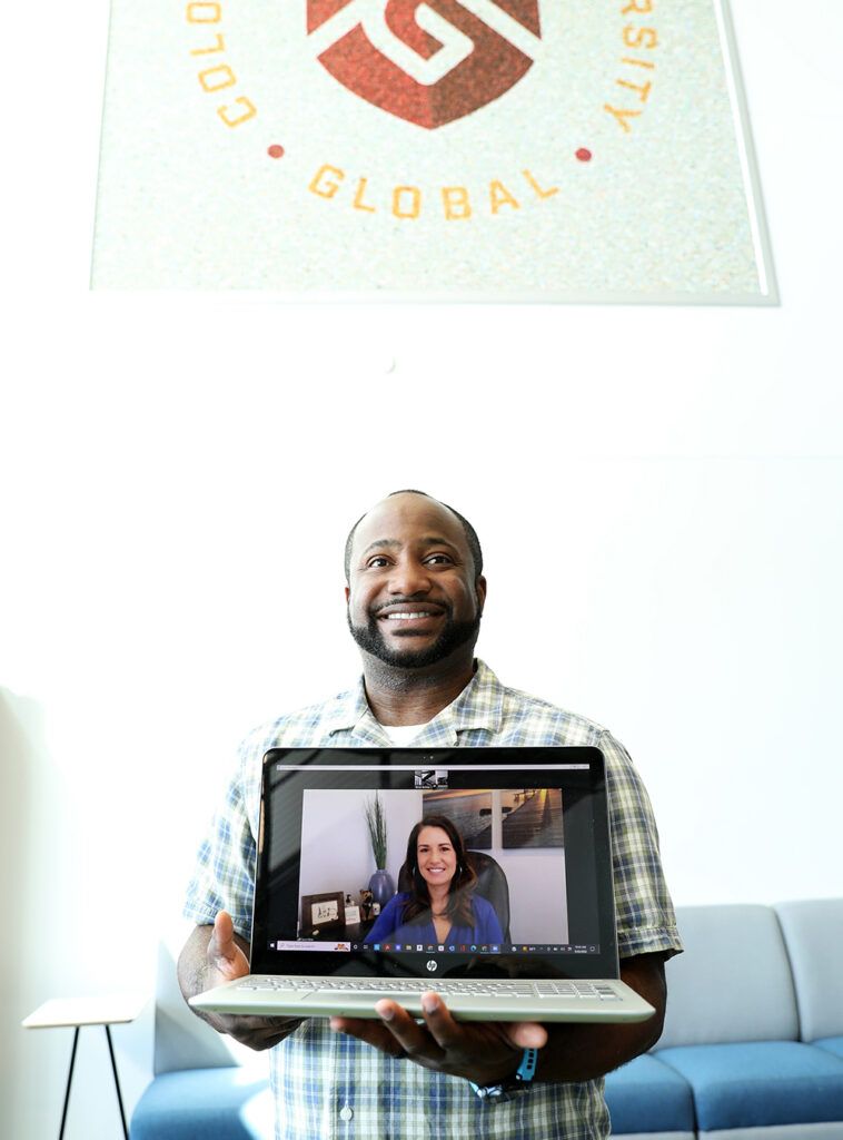 Man holds a laptop facing the camera with a woman on a video meeting on the screen.