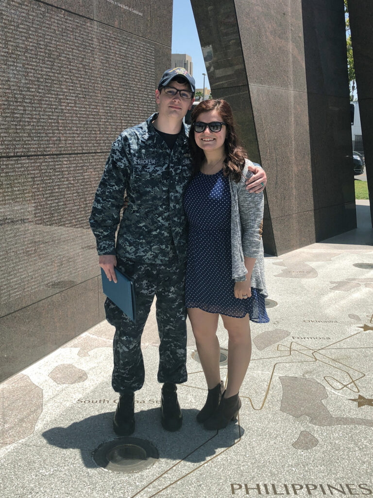 Two people, one in military fatigues, stand in front of a memorial.