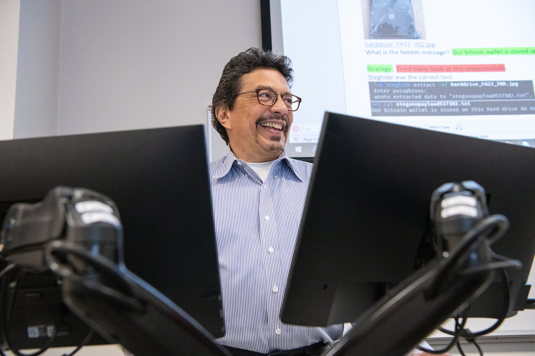 A person sits behind two computer monitors and smiles off camera.