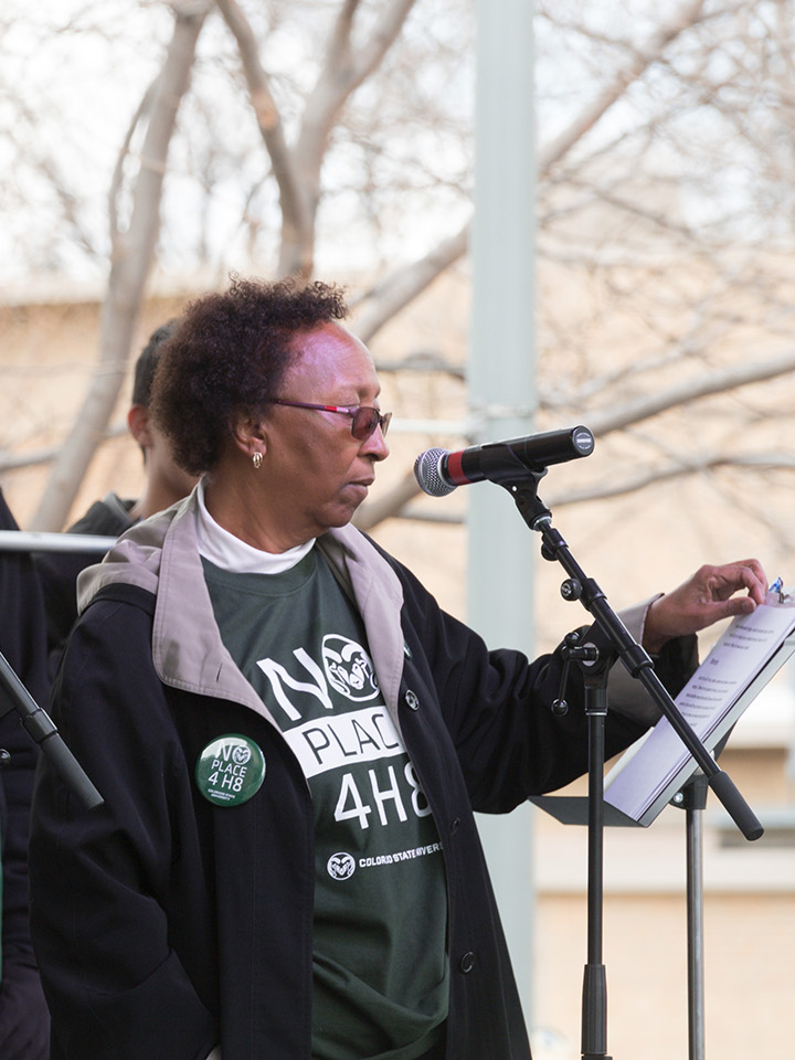 A woman in sunglasses and a CSU shirt stands at a microphone.
