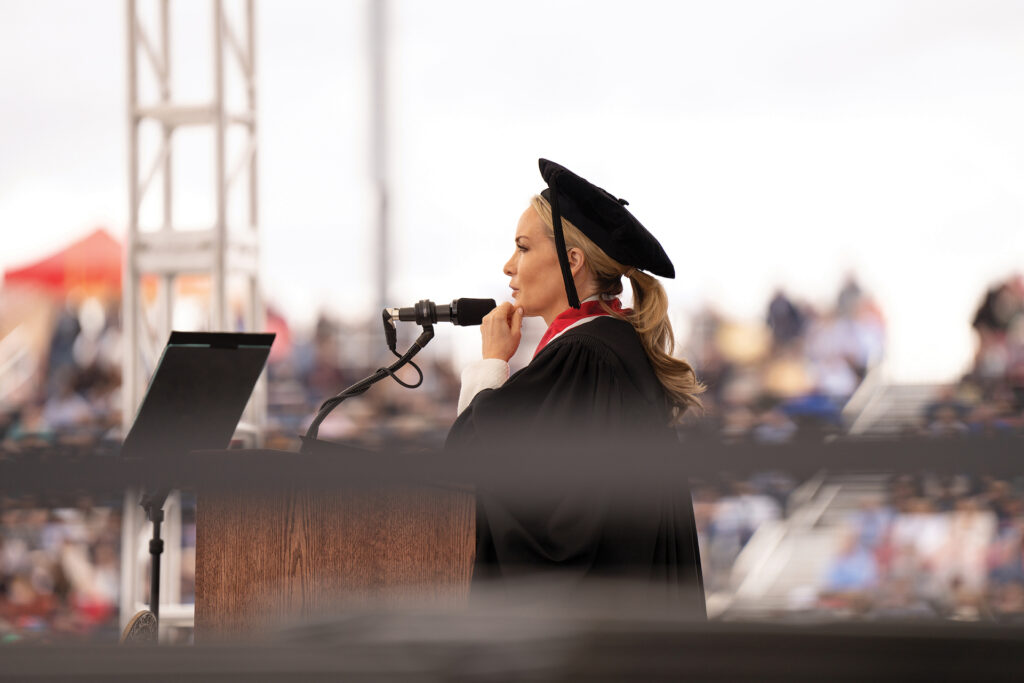 A speaker in graduation regalia stands at a podium and speaks into a microphone.