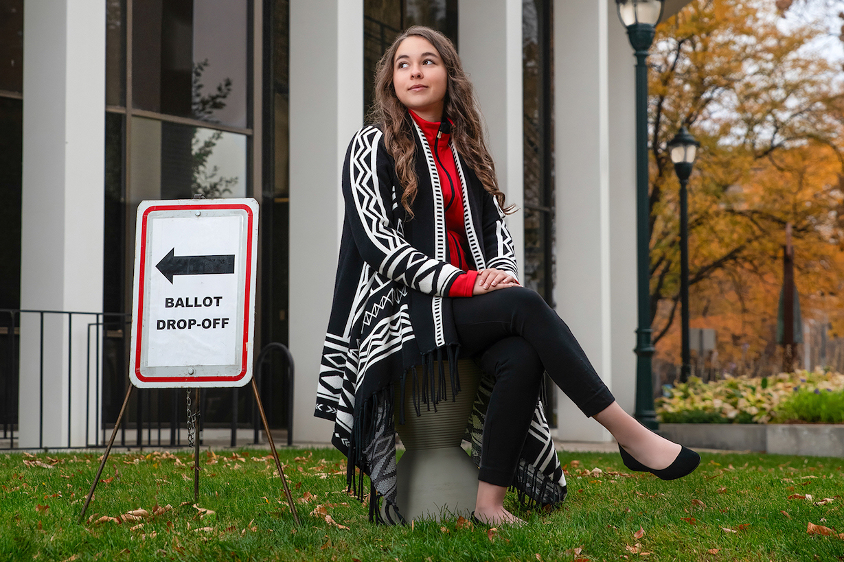 A woman poses in front of a building with a yard sign that reads "ballot drop off" with an arrow.