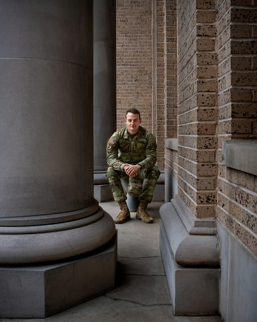 A man in military fatigues poses near large building columns.
