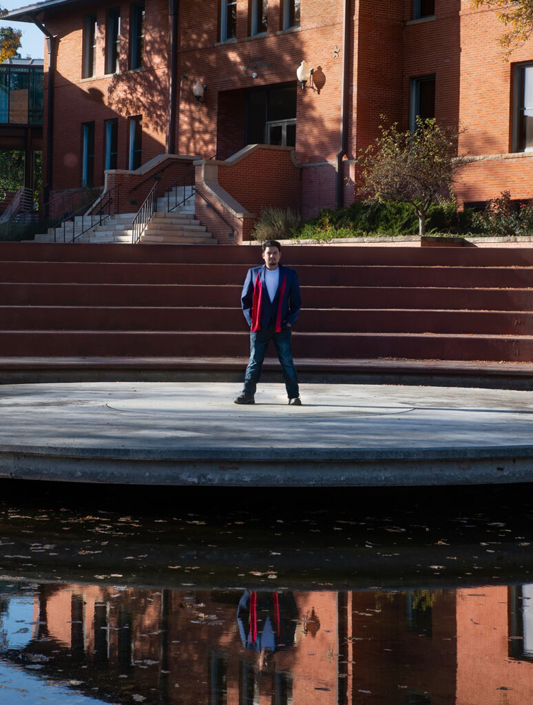 A man poses for a photo in front of a brick building with his reflection in a pool of water in the foreground.