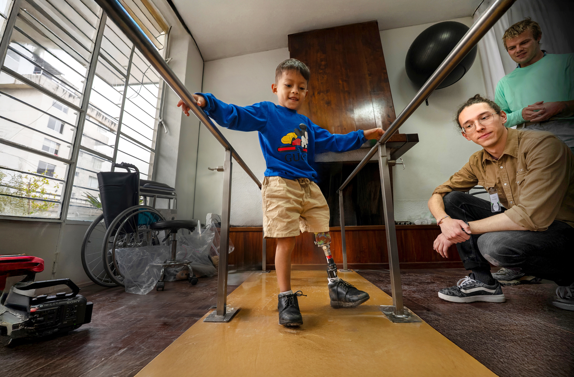 A young boy with a prosthetic leg walks in between two railings with two other people looking on.