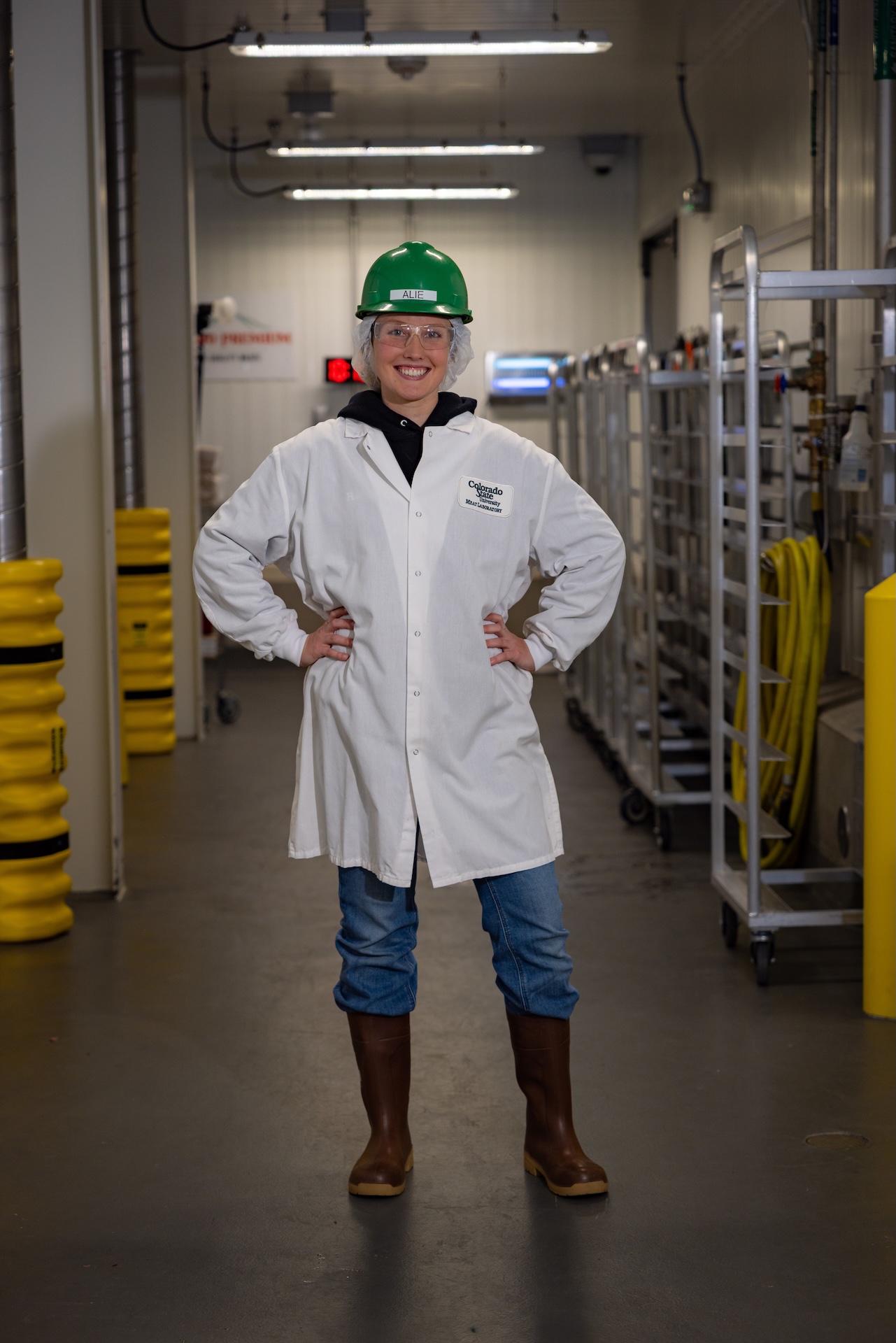 A woman poses in a lab coat, green hard hat, and knee-high rubber boots.