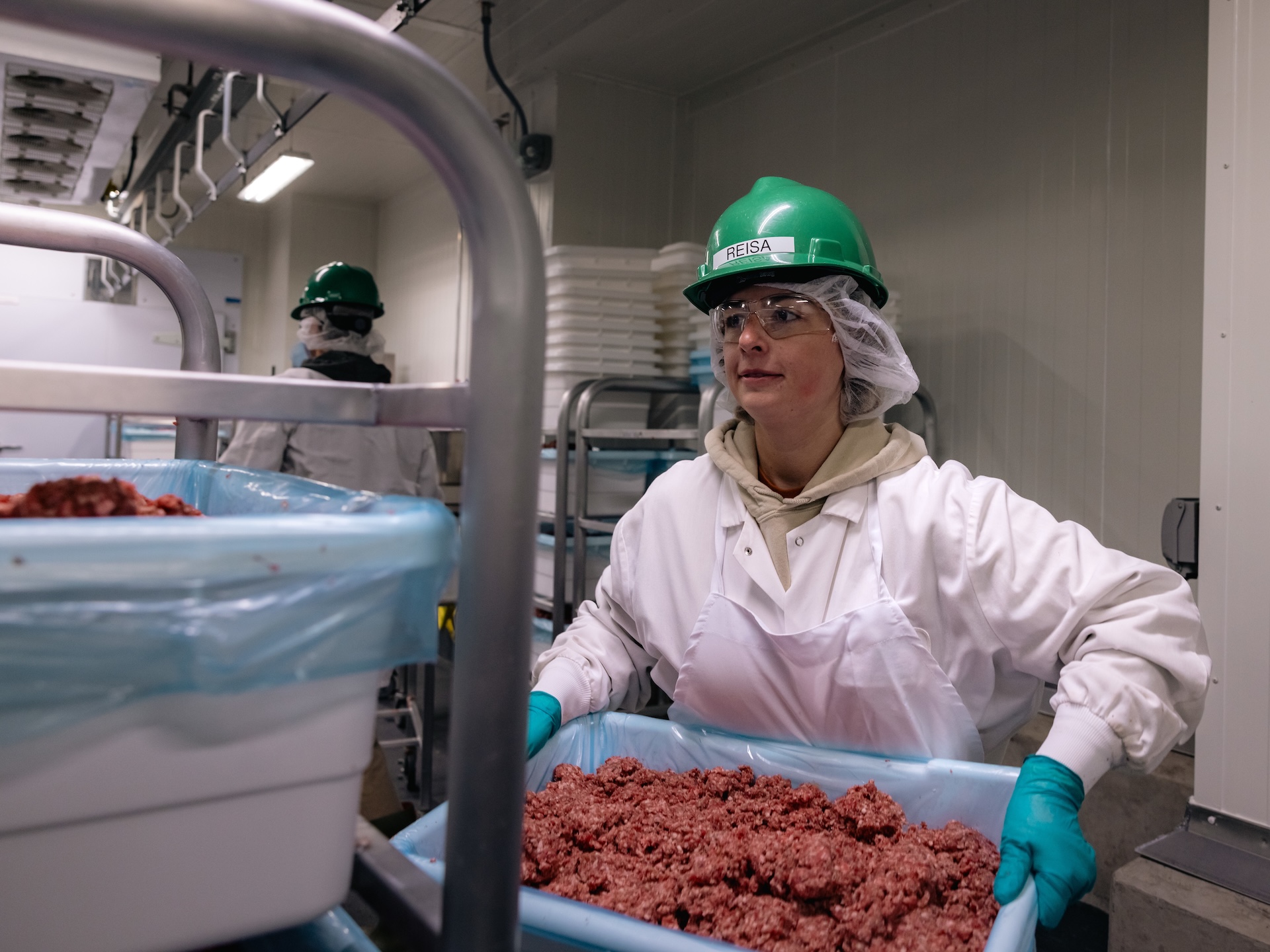 A woman in a green hard hat and safety goggles carries a tray of raw ground beef.