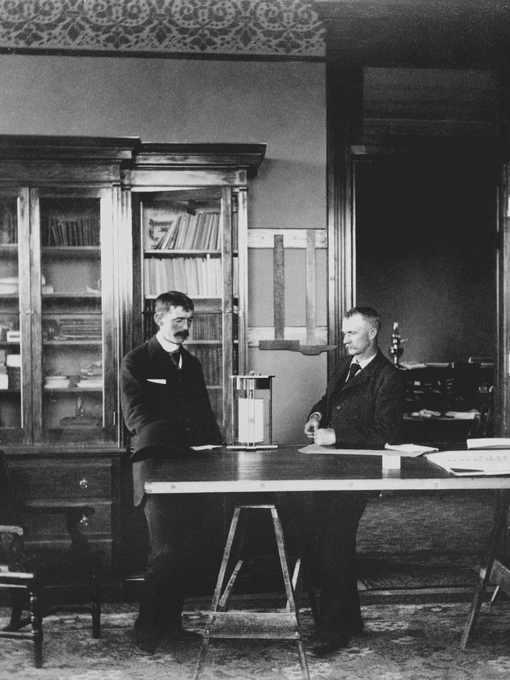 Historical photo of two men standing together at a table with a glass-fronted bookcase in the background.