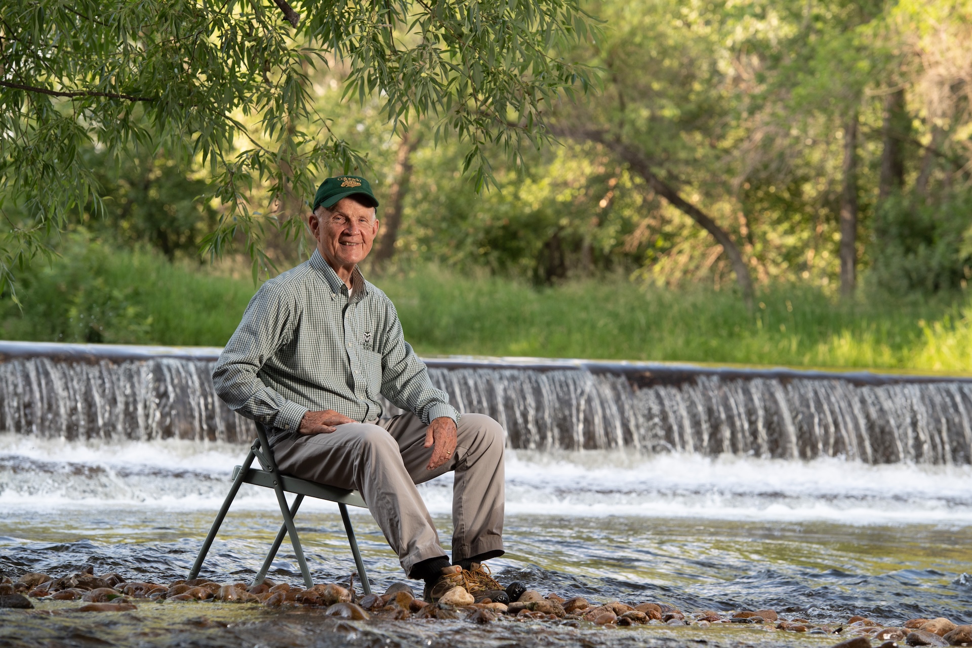 A man sits in a folding chair near a river and a short waterfall.