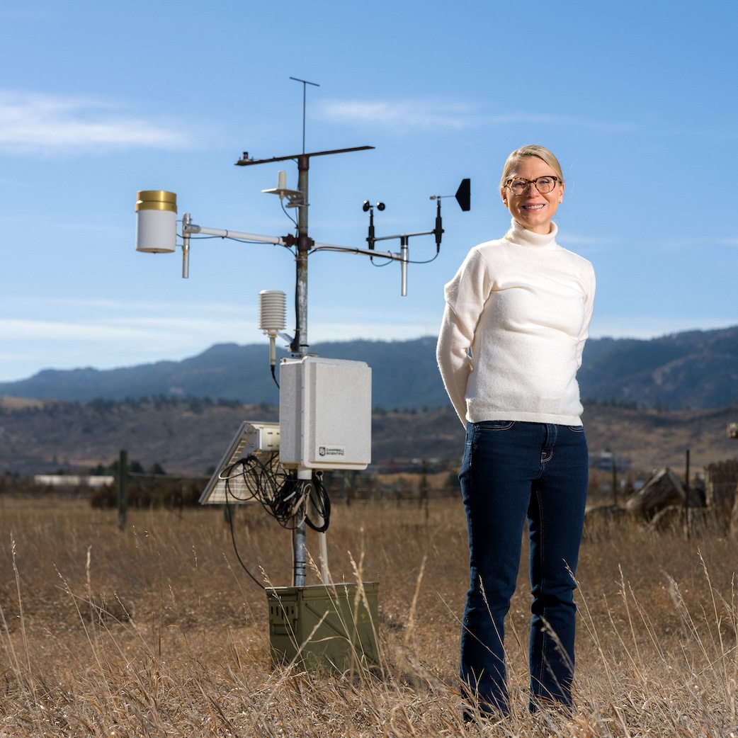 A woman in a white sweater stands in a field with a weather station in the background.