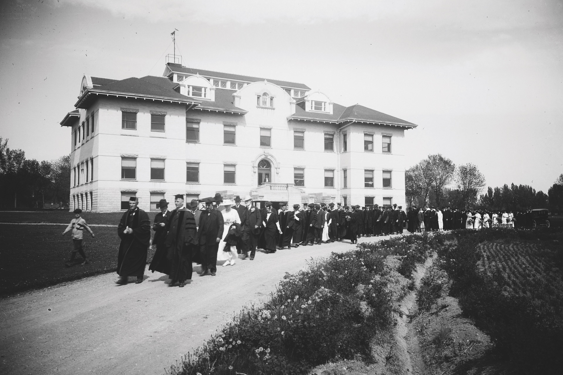 Historical photos of graduates in regalia lined up on a road outside of a white building.